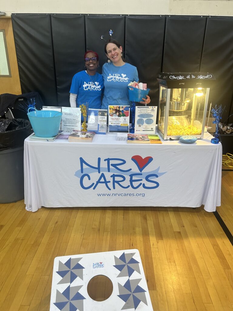 Two women in blue NRV Cares tshirts stand behind an NRV Cares table with a popcorn machine on it