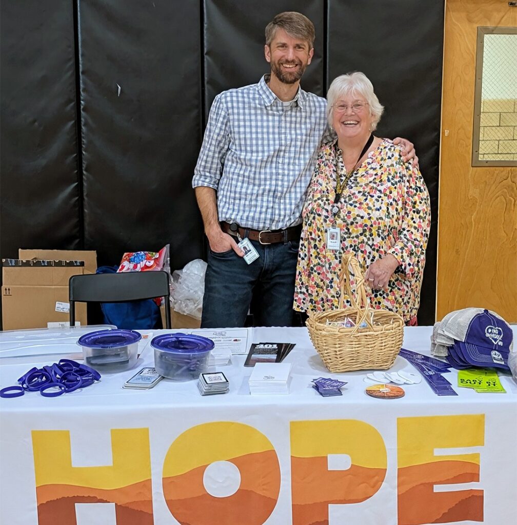 A man and a woman smile for the camera together behind a table with a tablecloth that reads "HOPE"