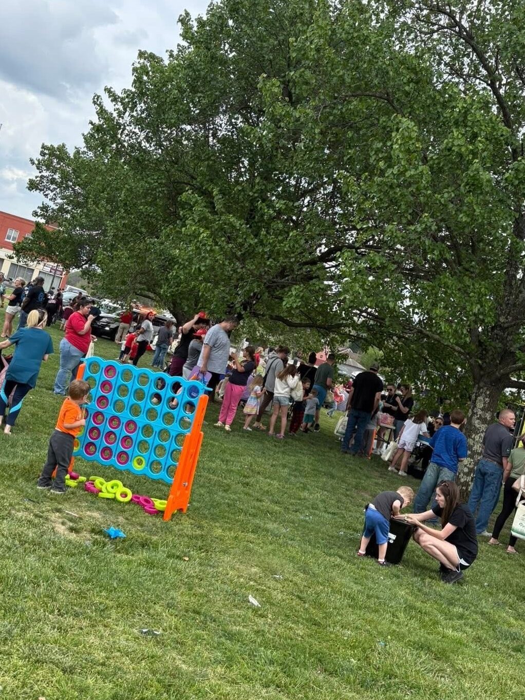 Families and children gather in the grass for an outdoor event; a child in an orange shirt plays a giant game of cornhole